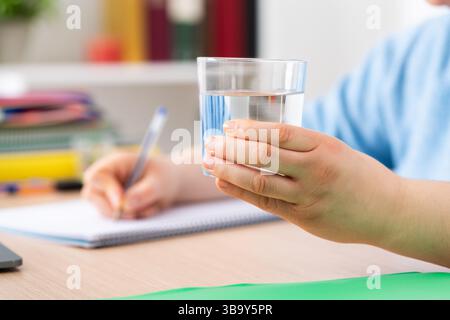 Nahaufnahme einer Studentenhand, die ein Glas Wasser hält und zu Hause mit Notizbuch studiert Stockfoto