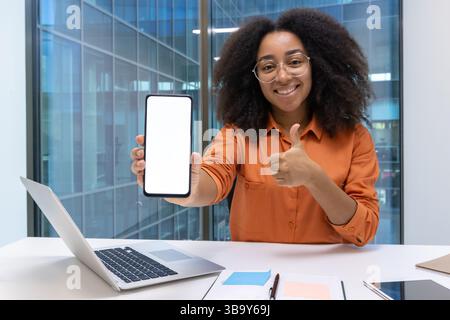 Eine fröhliche junge Frau mit Brille zeigt einen leeren Smartphone-Bildschirm und zeigt in ihrem Büro die Daumen nach oben. Stockfoto