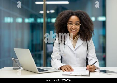 Eine freundliche und lächelnde schwarze Ärztin arbeitet an ihrem Schreibtisch. Sie trägt eine Brille und ein Stethoskop. Stockfoto