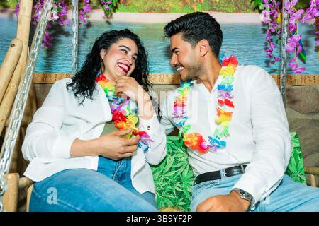 Glückliches Paar, das bunte hawaiianische Girlanden trägt, lächelt und ihre gemeinsame Zeit auf einer Schaukel genießt Stockfoto
