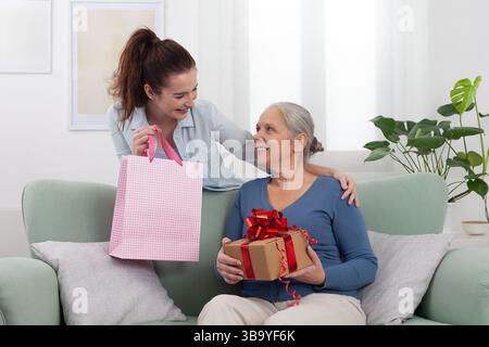 Glücklicher, generationsübergreifender Familienmoment zu Hause. Lächelnde junge Frau oder Enkelin, die ihrer fröhlichen älteren Mutter oder Großmutter ein Geschenk schenkt Stockfoto