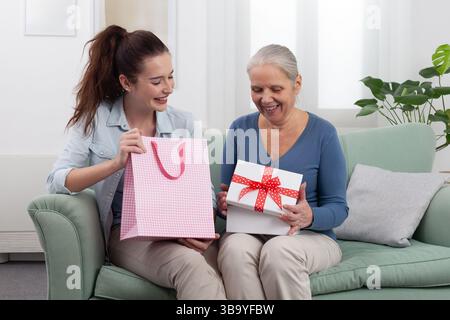 Glücklicher, generationsübergreifender Familienmoment zu Hause. Lächelnde junge Frau oder Enkelin, die ihrer fröhlichen älteren Mutter oder Großmutter ein Geschenk schenkt Stockfoto