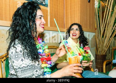Zwei fröhliche Frauen, die hawaiianische Leis tragen, treffen sich und trinken Cocktails in einer Tiki-Bar mit Bambusdekoration Stockfoto