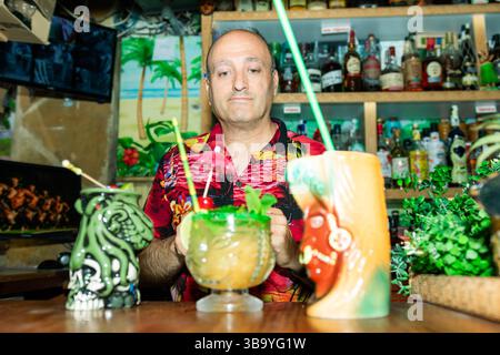 Der Barkeeper hält zwei tropische Cocktails in einer Tiki-Bar und trägt ein hawaiianisches Hemd Stockfoto