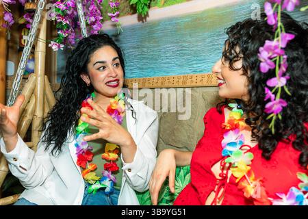Zwei Frauen mit farbenfrohen hawaiianischen Leis genießen Gespräche und Cocktails in einer tropischen Tiki-Bar oder einem Restaurant Stockfoto