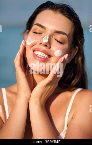 Beauty Woman wendet fröhlich Gesichtscreme am Strand an, um ihre Hautpflege Routine und die Bedeutung des Sonnenschutzes für gesunde Haut zu übernehmen. Haut C Stockfoto