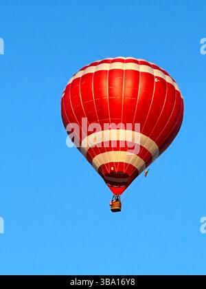 Rot-weißer Heißluftballon, der während der Cheshire Balloon Fiesta, Tattenhall, Großbritannien, am 6. August 2023 gegen einen klaren blauen Himmel schwingt. Stockfoto