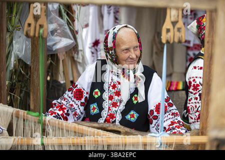 Eine alte belarussische oder ukrainische Frau in einem gestickten Hemd an einem Vintage-Webstuhl. Slawische ältere Frau in nationalen ethnischen Kleidern Stockfoto