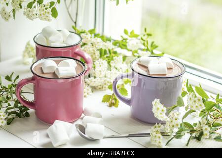 Heißer Kakao mit Marshmallows in rosafarbenen Bechern und frischen Frühlingsblumen auf der Fensterbank. Gemütliches Wohnkonzept. Geringe Schärfentiefe Stockfoto