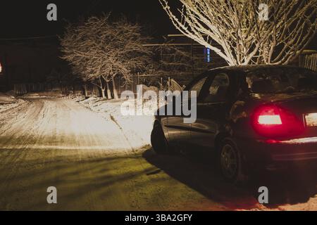 Die hellen Scheinwerfer eines schwarzen Autos beleuchten die schneebedeckte Straße im Winter Stockfoto