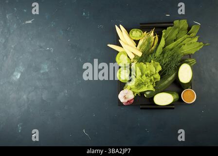 DAS GEMÜSE IN DER SCHACHTEL AUF DEM STEIN, EIN DUNKLER HINTERGRUND. JUNGE GRÜNE ZWIEBELN KNOBLAUCH ZUCCHINI HELLE GEWÜRZE LIEGEN IN EINEM HOLZTABLETT MIT GRIFFEN AUF EINEM DUNKLEN Stockfoto