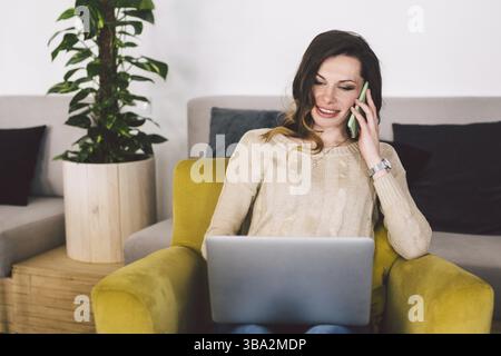 Glückliche, erfolgreiche Geschäftsfrau, die mit dem Handy anruft, während sie im Café sitzt, lächelnde Unternehmerin, die einen Laptop benutzt und mit ihr gesprochen hat Stockfoto