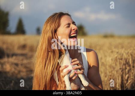 Young woman in wheat field lit by afternoon sun, holding puppy trying to pose, but dog is hidden behind hair, licking and chewing her ear, Liptovsky H Stockfoto