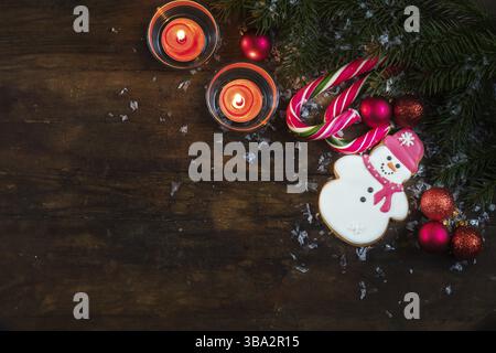 Weihnachten und Neujahr Komposition. Lebkuchenschneemänner, Tannenzweige, Kugeln, zwei Kerzen und Zuckerrohr mit Schnee auf dunklem Holzhintergrund Stockfoto