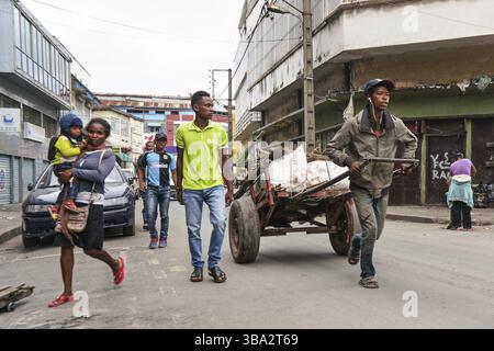 Antananarivo, Madagaskar - 24. April 2019: Unbekannter madagassischer Mann, der mit weißen Taschen den Wagen zieht, Frau mit Kind auf den Händen, die nahe kommt, mehr Einheimische Stockfoto