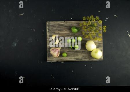 DAS GEMÜSE AUF DEM BRETT. ZWIEBELN KNOBLAUCH TOMATEN DILL AUF GRAUEM HOLZBRETT AUF EINER ERHÖHTEN STEINOBERFLÄCHE Stockfoto