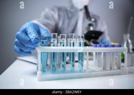 Wissenschaftler oder Sanitäter in Schutzkleidung, nimmt Reagenzglas mit blauem Reagenz, Laborglas mit chemischer Flüssigkeit, Mikroskop, Labor Stockfoto