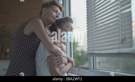 Erwachsene Tochter umarmt mit liebevoller älterer Mutter 90 Jahre in Maske stehen am Fenster, Verwandten aus verschiedenen Generationen, Frauen stehen im embrac Stockfoto