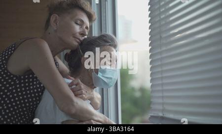 Erwachsene Tochter umarmt mit liebevoller älterer Mutter 90 Jahre in Maske stehen am Fenster, Verwandten aus verschiedenen Generationen, Frauen stehen im embrac Stockfoto