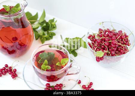 Rote Johannisbeere trinken in durchsichtiger Glaskaraffe und Tasse. Vase aus klarem Glas mit roten Johannisbeeren auf weißem Holzhintergrund Stockfoto