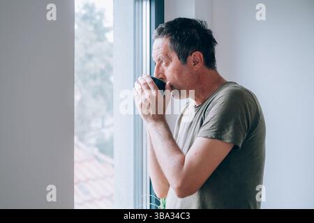 Nachdenklicher Mann, der morgens Kaffee am Fenster der Wohnung trinkt, während er hinausschaut und nachdenkt. Coffeindosis zum Tagesbeginn. Selektiv fo Stockfoto