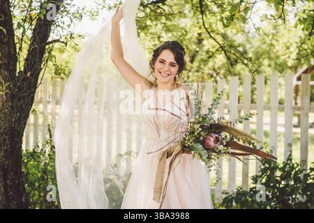 Schöne Braut mit Blumenstrauß Blumen steht auf Wald Hintergrund. Landhausstil. schöne Braut in zarten Kleid im Freien. Nahaufnahme, Porträt der jungen Stockfoto