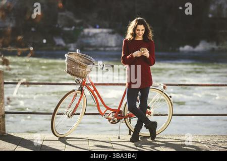 Thementourismus auf Fahrrad und moderne Technik. Schöne junge kaukasische Frau steht in der Nähe des roten Retro-Fahrrads am Rhein Basel Swiss WIN Stockfoto