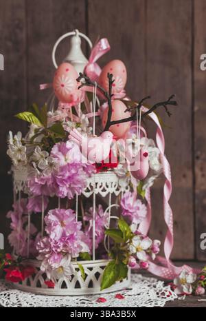 Frühlingsdekor: Rosafarbene Vögel auf dem Ast in einem schäbigen, schicken Käfig mit Blumen Stockfoto