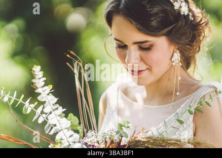 Schöne Braut mit Blumenstrauß Blumen steht auf Wald Hintergrund. Landhausstil. schöne Braut in zarten Kleid im Freien. Nahaufnahme, Porträt der jungen Stockfoto