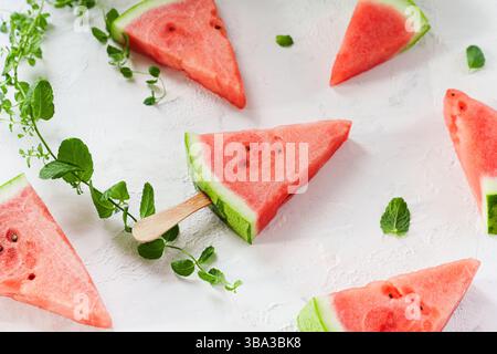 Eine dreieckige Wassermelonenscheibe mit Stab liegt auf weißem Hintergrund mit Minzblättern und Wassermelonenscheiben verstreut. Stockfoto
