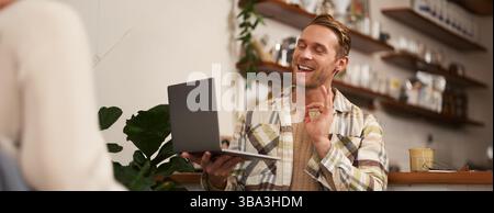 Porträt eines jungen lächelnden Mannes, ein Kollege nimmt an einem Online-Meeting vom Café Teil, Video-Chats auf Laptop, zeigt OK, Genehmigung von Hand eintragen, Kaffee trinken Stockfoto