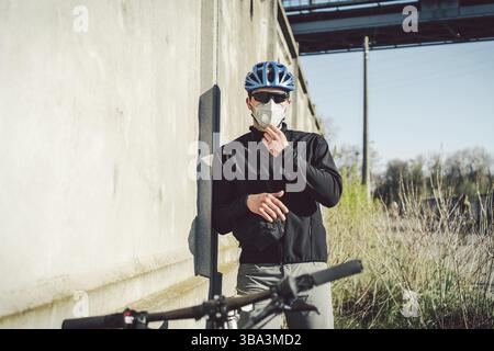 Ein Mann Radfahrer in einer schützenden Gesichtsmaske steht neben einem Fahrrad auf einem Hintergrund aus grauer Betonwand. Unkenntlich erkennbarer Kurier in Atemschutzmaske. Lieferung Stockfoto