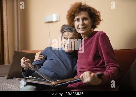 Seniorin und ihre Erwachsene Tochter schauen sich gemeinsam auf der Couch im Wohnzimmer ein Fotoalbum an und reden freudig über Erinnerungen. Wochenende mit Eltern Stockfoto