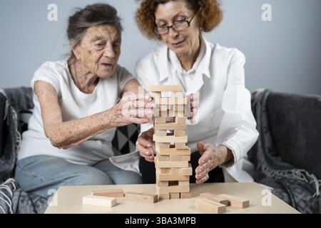 Ältere Frau spielt Jenga, baut einen Turm aus Blöcken. Älterer Arzt in weißem Laborkittel, Unterstützung für ältere Patienten, Entwicklung Logik, Feinmotorik, EIN Stockfoto
