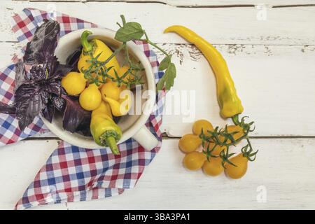 Gelber heißer Pfeffer und gelbe Tomaten mit violettem Basilikum in einem weißen Keramiktopf auf einem karierten Tuch. Draufsicht. Kopierbereich Stockfoto