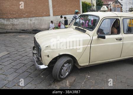 Antananarivo, Madagaskar - 24. April 2019: Weißes Taxiauto, das langsam über Fliesensteinstraße fährt, Gruppe unbekannter madagassischer Menschen im Hintergrund. Madagas Stockfoto