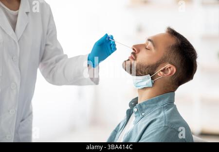 Coronavirus-Testverfahren. Doktor, der nasalen PCR-Test für Millennial Kerl macht, mit Abstrichbesteck zu Hause Stockfoto