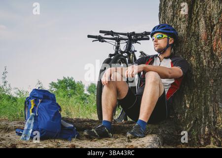 Ein männlicher Mountainbiker, der auf einer Radtour sitzt und mit seinem Mountainbike auf dem Boden unter einem Baum sitzt, steht neben ihm und genießt die wunderschöne Natur aro Stockfoto