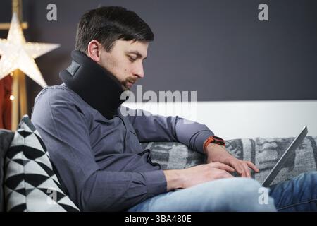 Mann Mitarbeiter nach Unfall zu Hause arbeiten hinter Laptop und fühlt Schmerzen im Nacken, verwendet schwarzen Kragen Unterstützung. Männlich mit Nackenverletzung freiberuflich prog Stockfoto