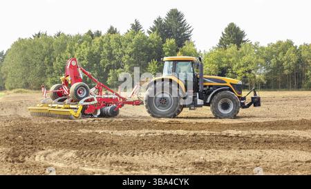 Gelber Traktor zieht roten Sämechanismus über trockenes Feld, Bäume im Hintergrund, Liptovsky Hradok, Slowakei, Europa Stockfoto