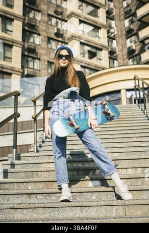 Urbane Frau mit Skate. Hipper-Mädchen mit Skateboard in der Stadt. Konzept für Extremsport und Emotionen. Alternativer Lebensstil. Modische Hüftmädchenhaltung Stockfoto