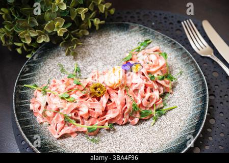 Rosafarbenes Gourmet-Pastagericht mit Fettuccine mit cremiger Sauce, Garnelen, essbaren Blumen und frischen Kräutern, serviert auf einer modernen Keramikplatte. Stockfoto