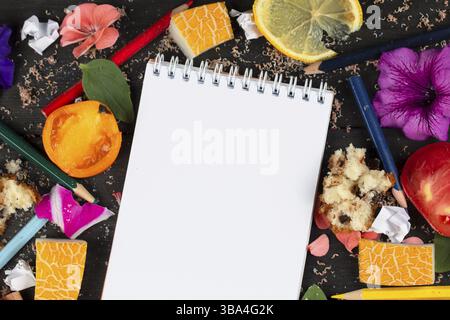 Papiernotizbuch mit leeren Seiten, zerknitterten Papierkugeln und Buntstiften, verstreuten Lebensmitteln auf Holzhintergrund, kreatives Kochrezept-Schreibkonzept. Kommi Stockfoto