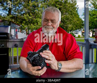 Ein älterer Mann mit Bart sitzt auf einer Veranda und inspiziert seine Kamera sorgfältig. Die Sonne scheint auf einem gut gepflegten Hof in einer ruhigen Vorstadt-Umgebung, die ein Gefühl der Freizeit weckt. Stockfoto