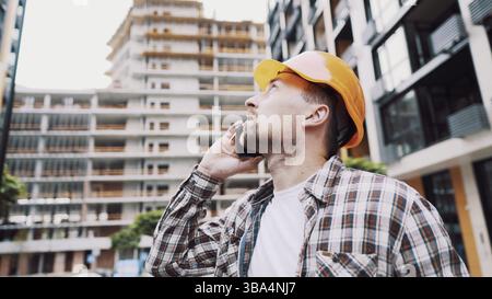 Porträt eines jungen Handwerkers, der Anrufe macht, während er auf der Baustelle steht. Ein Techniker, der auf einer Baustelle telefoniert. Builder in Helm Co Stockfoto