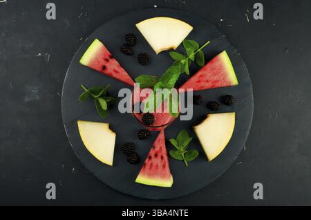 MELONE WASSERMELONE BROMBEEREN MINZE UND EIN GLAS SAFT AUF RUNDEM HÖLZERNEN HINTERGRUND. REIFE WASSERMELONE SAFTIGE MELONE BROMBEERMINZCOCKTAILS AUF EINEM DUNKLEN WO Stockfoto