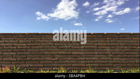 Ziegelmauer mit weißen Wolken am blauen Himmel. Blauer Himmel über der roten Ziegelmauer. Sträucher und Ziegelzaun auf blauem Himmel Hintergrund Stockfoto