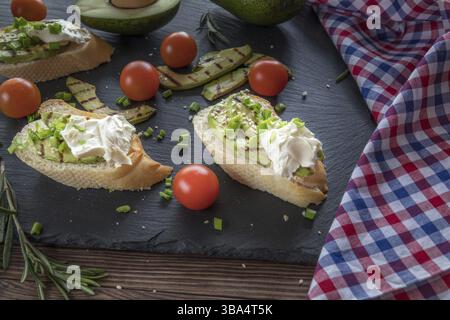 Brot mit gegrillten Avocadoscheiben und Frischkäse auf einem schwarzen Steinbrett. Frische Petersilie und Rosmarin. Vegetarisches, veganes Konzept Stockfoto