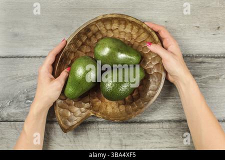 Blick auf die Tischplatte auf grauem Holztisch, Frauenhände halten eine hölzerne Schüssel mit drei ganzen grünen Avocados, Liptovsky Hradok, Slowakei, Europa Stockfoto