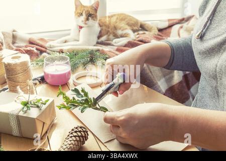 Frau wickelt umweltfreundliche Weihnachtsgeschenke mit braunem Papier, Schnur und natürlichen Zweigen und Dekoelementen zu Hause auf Holztisch ein Stockfoto